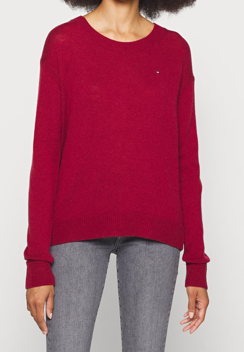 Femme portant un pull rouge à manches longues en tricot avec poignets et ourlet côtelés, associé à un jean gris ajusté.