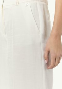 Jasno beżowe, dopasowane spodnie wykonane z fakturowanej tkaniny, z bocznymi kieszeniami i subtelnym połyskiem. Zbliżenie na pasek i dłoń.