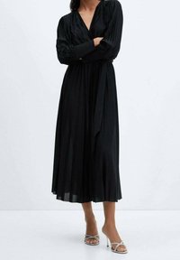 Robe noire plissée à manches longues et décolleté en V profond, cintrée à la taille. Portée avec des sandales à talons aiguilles argentées.