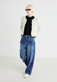 Cardigan alb tricotat cu țesătură deschisă, cu margini negre și nasturi; top negru; blugi din denim cu croială largă și embellismente florale; adidași albi.