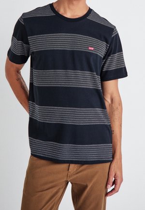 Homme portant une chemise à manches courtes à rayures horizontales noires et blanches avec un logo Levi's rouge, et un pantalon marron, tatouages visibles sur le bras.