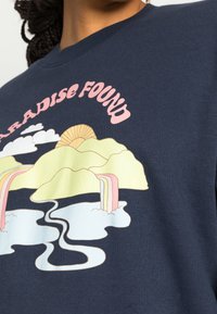 Námořnický svetr s grafickým designem obsahujícím hory, vodopády a slunce. Text zní "PARADISE FOUND" v růžové barvě. Měkký, bavlněný materiál.