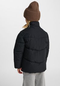 Svart puffjacka med hög krage och horisontellt kviltad design, med en matt finish och avslappnad passform, parat med en brun ribbad mössa.