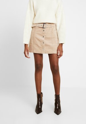 Vrouw die een beige ribfluwelen minirok met knopen en riem draagt, een crèmekleurige gebreide trui en donkerbruine textuurschoenen tot boven de enkel, staand op een witte achtergrond.