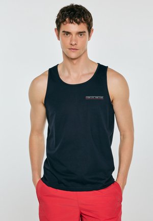 Mężczyzna stojący z rękami w kieszeniach, ubrany w czarny tank top z napisem i jaskrawoczerwone krótkie spodenki na jednolitym tle.