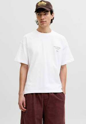Jeune homme portant un T-shirt blanc avec un petit texte sur la poitrine et un pantalon en velours côtelé marron, portant également une casquette marron foncé avec un motif jaune.