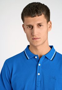 Niebieska koszulka polo wykonana z teksturowanej tkaniny, z kołnierzykiem w kontrastowe niebieskie i czarne paski, trzema guzikami oraz logo na piersi.