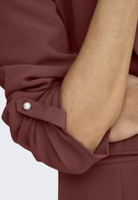 Bordo blůza s ohrnutými rukávy, s knoflíkovým zapínáním a perlovým detailem. Hladká, strukturovaná tkanina ukazuje uvolněný střih.