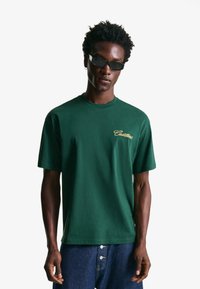 Green cotton t-shirt with a crew neck, short sleeves, and a gold "Cadillac" logo on the left chest. Worn with denim pants.