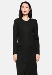 Langes schwarzes Kleid mit langen Ärmeln, rundem Ausschnitt, vorderer Knopfleiste, zwei vorderen Taschen und geradem Schnitt. Weiche Stoffstruktur.