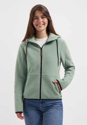 Smiling woman wearing light green zip-up hoodie with black drawstrings and blue jeans, standing against plain white background.