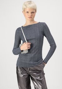 Szürke bordázott, hosszú ujjú felső, széles nyakkivágással, fémes ezüst nadrággal párosítva. Egy kis, textúrált ezüst táska van nála.