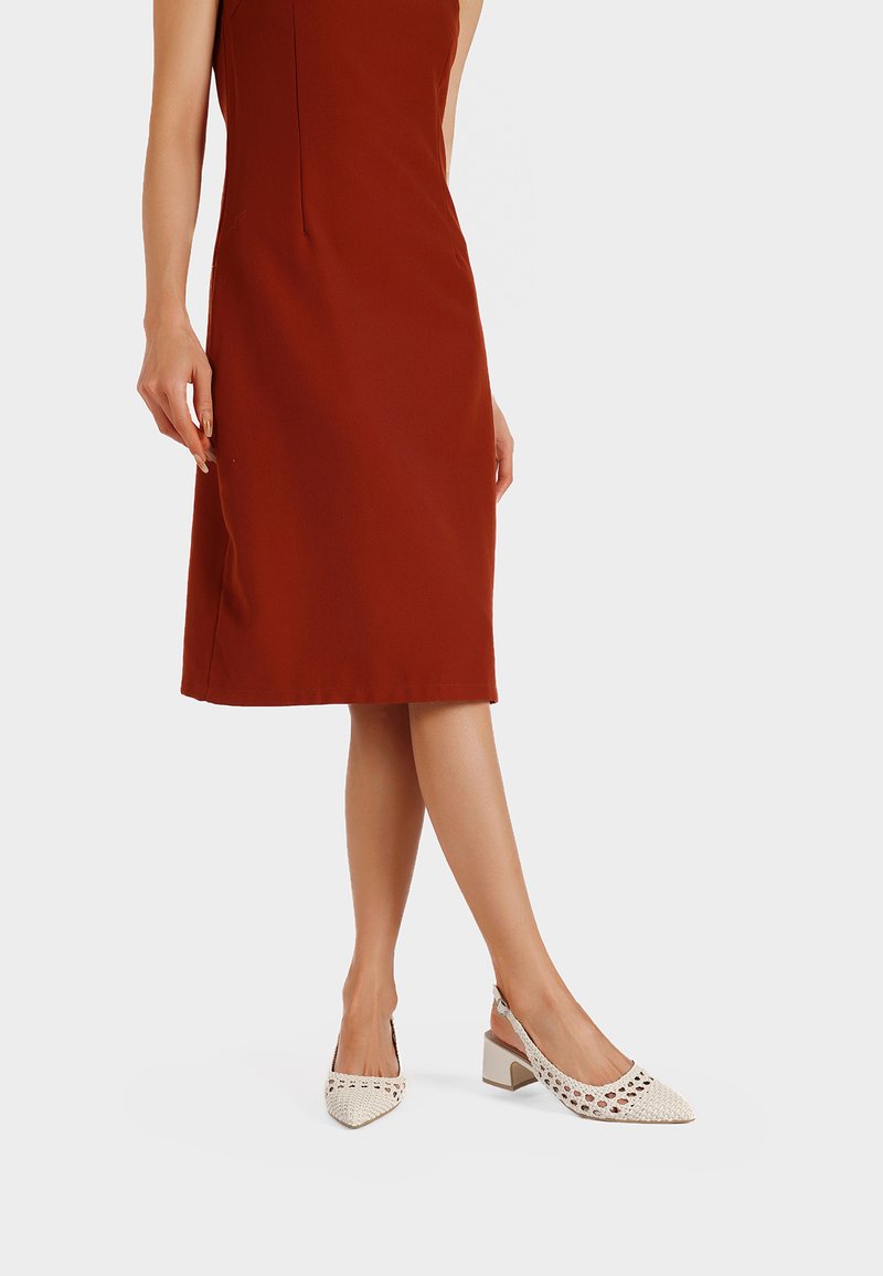 Scarpe con tacco alto beige intrecciate, con punta a punta e design ritagliato, abbinate a un vestito senza maniche color ruggine.