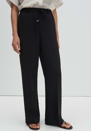 Persona che indossa pantaloni neri larghi con coulisse e piega frontale, abbinati a infradito nere e una maglietta bianca a righe infilata dentro.