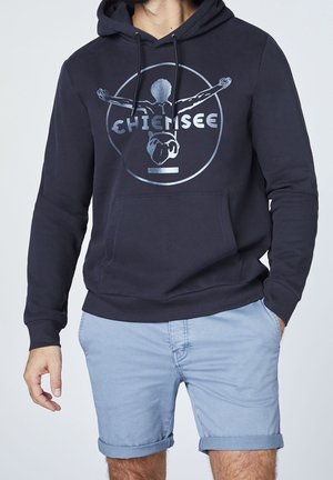 Felpa blu navy con tasca frontale, decorata con una stampa grafica argentata e cappuccio con cordino. Abbinata a pantaloni corti arrotolati azzurri.