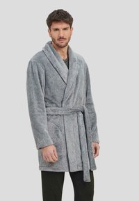 Bata de baño de algodón texturizado gris, con cuello de chal, Cintura ceñida con cinturón y dos bolsillos laterales, llevada sobre pantalones oscuros.