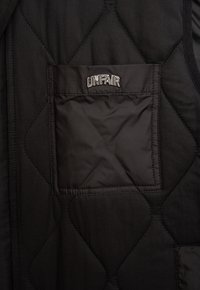 Svart kviltad väst med en framträdande fyrkantig ficka med det broderade ordet "UNFAIR." Slät textur med geometrisk sömnadsdetalj.