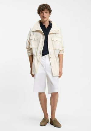 Nuori kiharatukkainen mies, joka pukeutuneena kermanväriseen takkiin mustan paidan päällä, valkoiset polvipituiset shortsit ja ruskeat loafersit, seisoo suorassa asennossa.