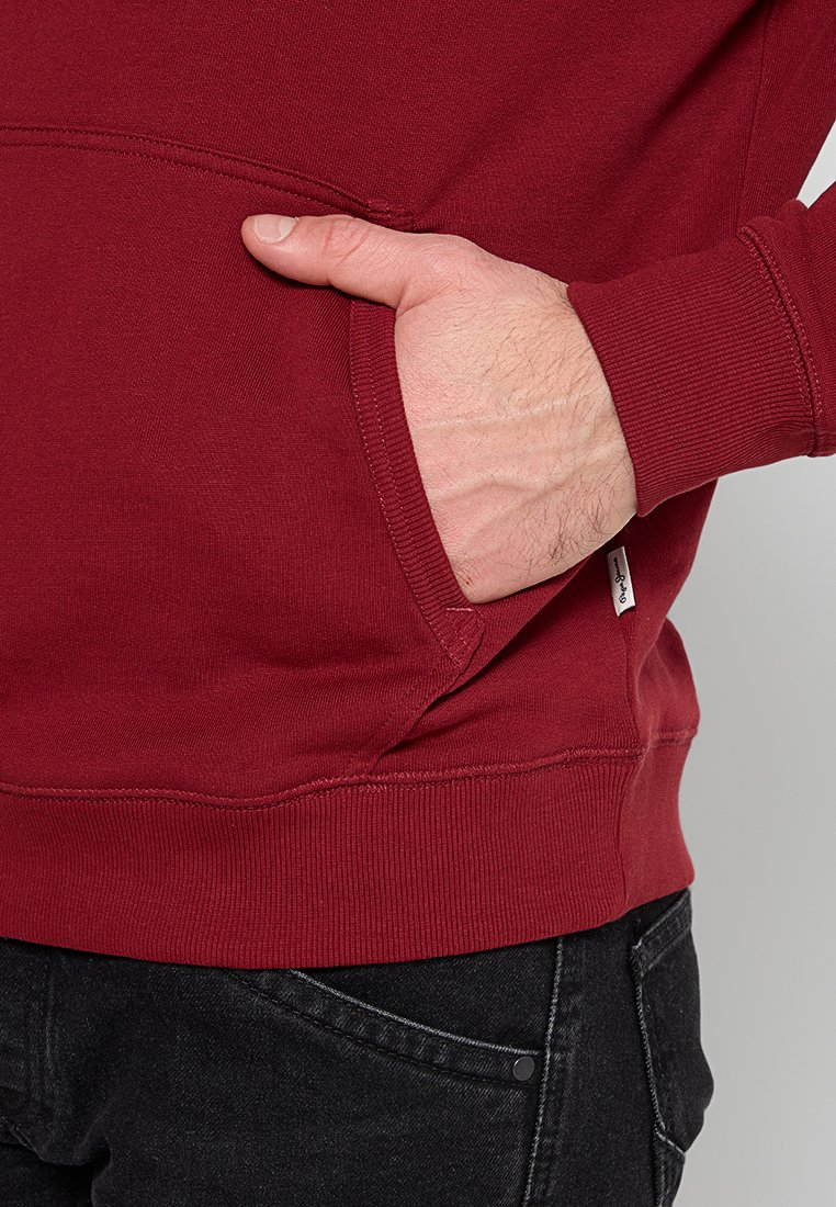 Sudadera burdeos con un bolsillo tipo canguro, puños acanalados y dobladillo. Material suave, con un detalle de etiqueta lateral, mostrada usada con jeans negros.