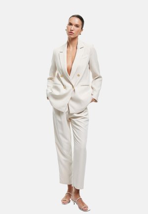 Traje blanco a medida con blazer de doble botonadura y pantalones de pierna recta, con botones dorados y escote en V profundo. Sandalias sencillas.
