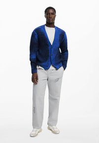 Cárdigan de punto azul con escote en V, cierre de botones y puños acanalados, combinado con pantalones gris claro con un estampado gráfico y zapatillas blancas.