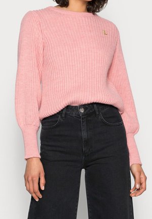 Femme portant un pull rose côtelé à manches longues bouffantes et un jean noir taille haute, raccourci à mi-cuisse.