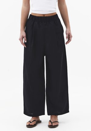 Person iført løstsiddende sorte bukser med høj talje, hvid kort tanktop og sorte klipklapper, stående mod en ensfarvet hvid baggrund.