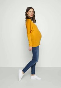 Top de maternidade em amarelo mostarda, corte solto com mangas longas, combinado com jeans skinny azul escuro e ténis brancos. Design simples, sem padrões notáveis.