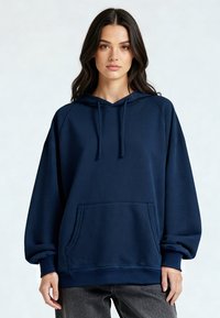 Jeune femme aux longs cheveux bruns portant un sweat à capuche surdimensionné bleu marine et un pantalon gris foncé, debout devant un fond uni.