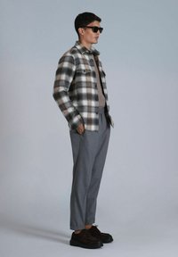 Karierter Flanellhemd in Schwarz, Weiß und Braun; brauner Strickpullover; graue Hose; dunkle Schuhe; Modell trägt Sonnenbrille.