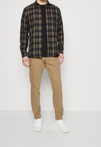 Camicia di flanella a quadri in nero, blu navy e senape; abbinata a pantaloni beige e sneakers bianche. Presenta bottoni e un colletto classico.