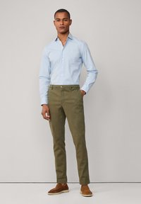 Lichtblauwe button-down shirt, olijfgroene chinos en bruine suède schoenen. Het shirt heeft lange mouwen; de broek is recht gesneden met een knoop aan de voorkant.
