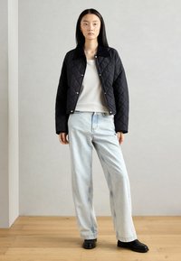 Veste matelassée noire avec col, boutons-pression argentés, portée sur un top blanc et un jean bleu clair. Des chaussures noires complètent la tenue.