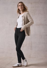 Cardigan a maglia color beige chiaro con maniche lunghe, bottoni sul davanti, abbinato a una t-shirt bianca, jeans skinny neri e sneakers bianche con dettagli neri.