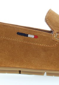 Tan semišová slip-on obuv s viditelným šitím a gumovou podrážkou. Vyznačuje se vyšívaným akcentem v modré, bílé a červené barvě poblíž vrchní části.