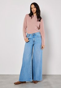 Hellrosa gerippter Pullover mit langen Ärmeln, der ein seitliches Muster aufweist, kombiniert mit hellblauen weit geschnittenen Jeans und braunen flachen Schuhen.