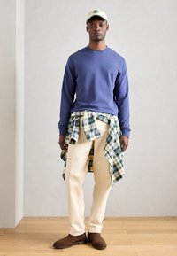 Mornarska pulover s majhnim logotipom, krema kariran plašč povezan okoli pasu, krema hlaci in rjavi gležnjarji na leseni tla.