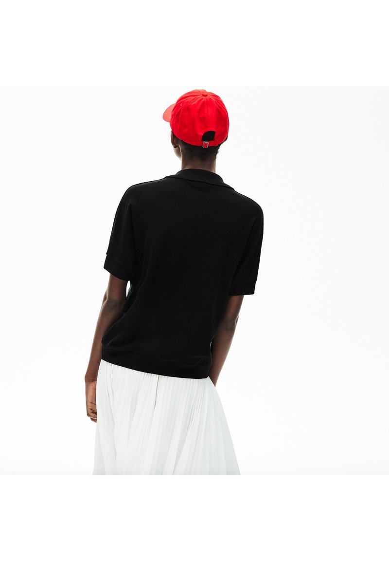 Schwarzes Kurzarm-Polohemd mit lässigem Schnitt, kombiniert mit einem weißen Faltenrock; rote Mütze mit verstellbarem Hinterriemen.