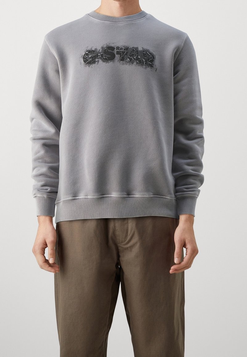 Personne portant un sweat-shirt gris avec le logo "G-STAR" et un pantalon marron, debout devant un fond clair uni.