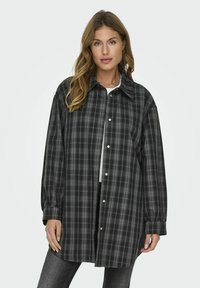 Camisa de franela de cuadros en negro y gris, de corte oversize, con cuello puntiagudo y cierre de botones en la parte delantera. Mangas largas con puños abotonados.