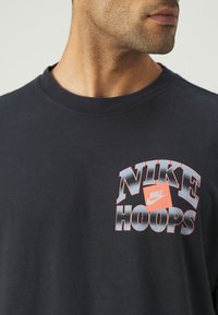 Czarna bawełniana koszulka z grafiką "NIKE HOOPS" w kolorze czerwonym i szarym. Okrągły dekolt, krótki rękaw i miękka faktura.