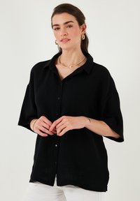 Black button-up shirt with a textured fabric, short sleeves, a collar, and a loose fit. Paired with white pants and gold jewelry accents.