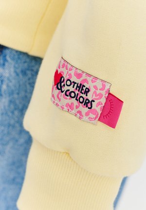 Żółta tkanina bluzy z prążkowanymi mankietami, z różową metką w panterkę oznaczoną napisem "Inne kolory" i małą grafiką oka.