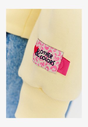 Żółta tkanina bluzy z prążkowanymi mankietami, z różową metką w panterkę oznaczoną napisem "Inne kolory" i małą grafiką oka.