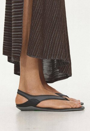 Piedi nudi con sandali slingback neri con sottili cinturini, in piedi su una superficie riflettente sotto una gonna a righe alla caviglia.