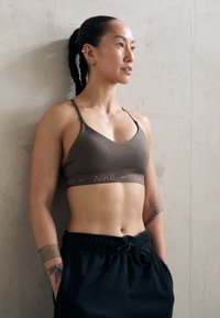 Sportos nő, aki fonott hajú, barna Nike sportmelltartót és fekete nadrágot visel, egy betonfal előtt áll, és oldalra néz.