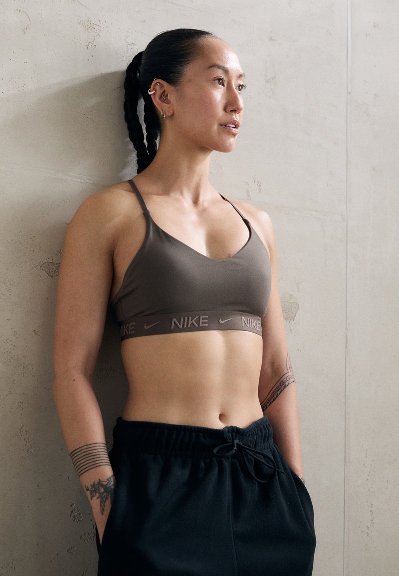 Sportos nő, aki fonott hajú, barna Nike sportmelltartót és fekete nadrágot visel, egy betonfal előtt áll, és oldalra néz.