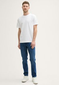 Camiseta de algodón blanca, de manga corta, combinada con jeans de mezclilla azul. El modelo lleva zapatillas blancas. Un conjunto limpio y casual en general.