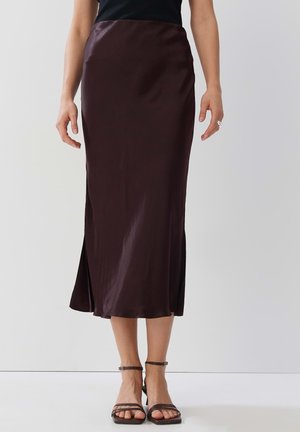 Dunkelbrauner Satinrock mit gerader Silhouette und seitlichem Schlitz. Begleitet von Riemchen-Heels, die die schlichte und glatte Textur unterstreichen.