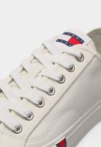 Baskets en toile blanche avec lacets blancs et œillets métalliques, arborant le logo Tommy Jeans sur la languette et près de la semelle.